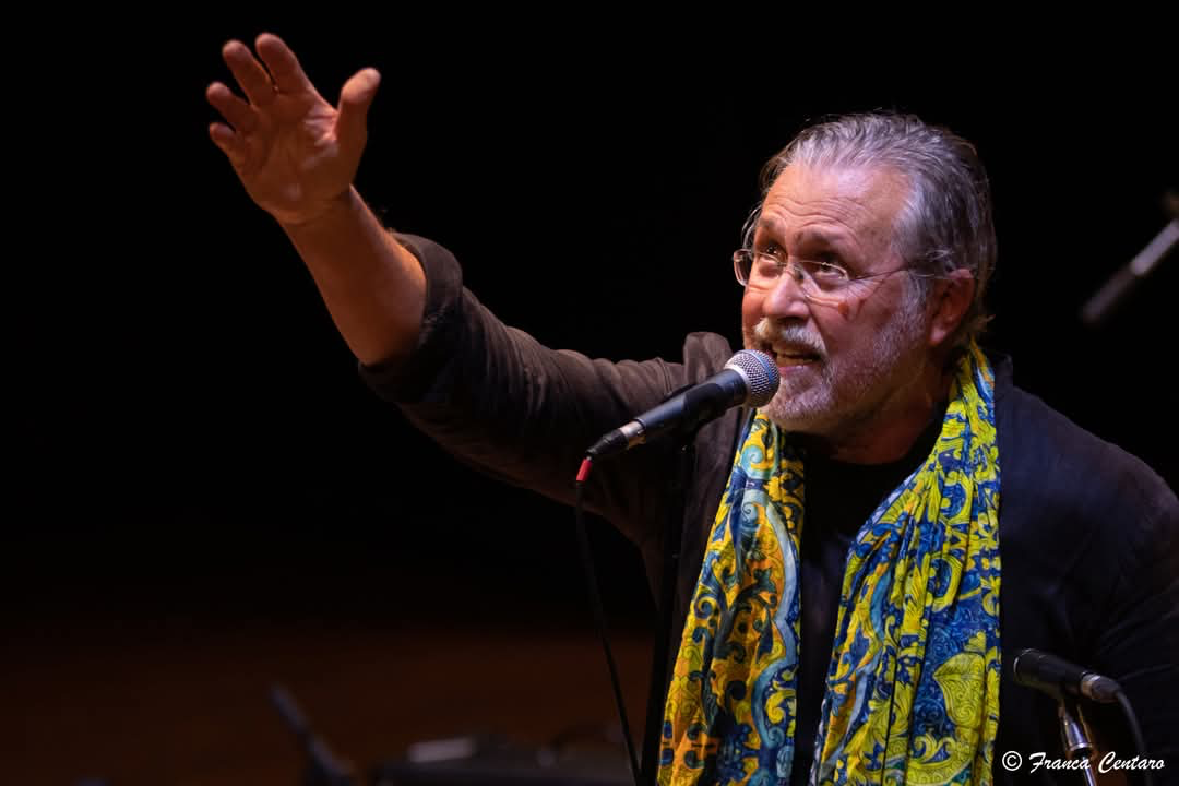 Pietro Romano canta al microfono con una sciarpa colorata e il braccio alzato
